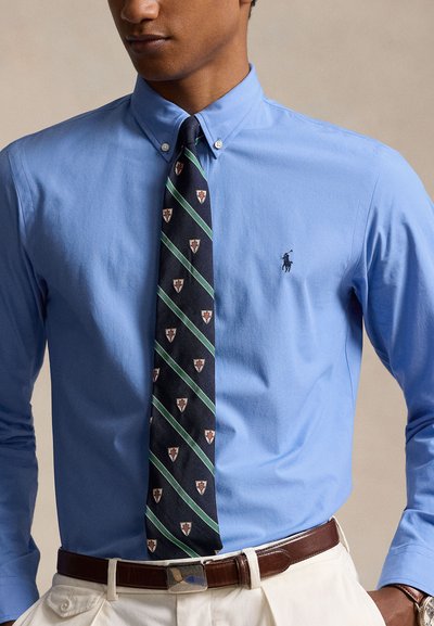 Ανοιχτό μπλε πουκάμισο με μικρό λογότυπο, σε συνδυασμό με σκούρα γραβάτα που έχει διαγώνιες πράσινες ρίγες και χαρακτηριστικά διακοσμητικά στοιχεία.