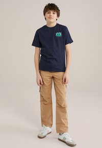 Marineblauwe t-shirt met korte mouwen en een kleurrijk grafisch zakontwerp, gecombineerd met beige cargobroek met zijzakken en witte sneakers.