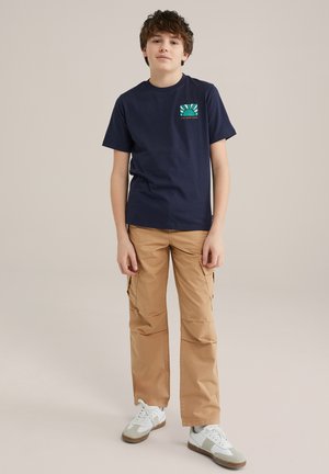 T-shirt bleu marine à manches courtes avec un design de poche graphique coloré, accompagné de pantalons cargo beiges avec poches latérales et de baskets blanches.