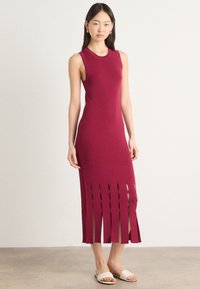 Dopasowana bezrękawnikowa sukienka w burgundowym dzianinie, z pionowymi wycięciami na końcu. Połączona z jasnymi sandałami wsuwanymi.