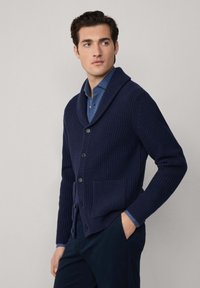 Cardigan blu navy con coste, colletto a scialle, quattro bottoni e tasca anteriore. Indossato sopra una camicia azzurro chiaro con bottoni e pantaloni navy.