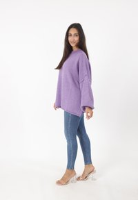 Lila Übergröße-Pullover mit Rundhalsausschnitt und abgesenkten Schulternähten, kombiniert mit eng anliegenden blauen Jeans und durchsichtigen Absatzsandalen.