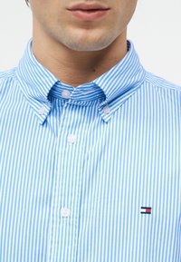 Άνδρας που φοράει μπλε και λευκό κάθετα ριγέ πουκάμισο με κουμπιά και γιακά με κουμπιά και ένα μικρό ορθογώνιο λογότυπο στο στήθος.