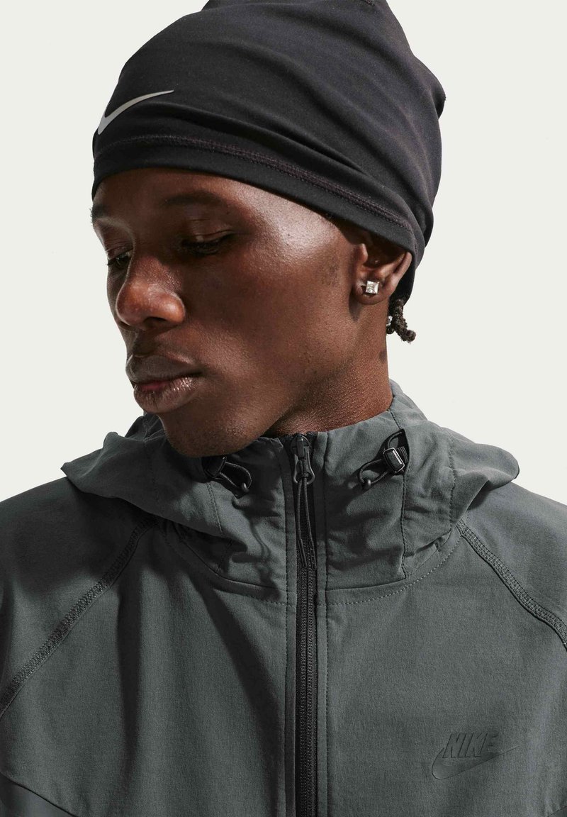 Man wearing a black Nike beanie, dark gray Nike zip-up jacket with hood, and square stud earrings, looking down with a neutral expression.