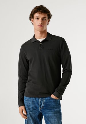 Giovane uomo con capelli ricci che indossa una polo nera a maniche lunghe e jeans blu, in piedi con una mano in tasca di fronte a uno sfondo neutro.