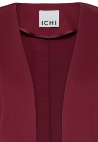 Gros plan sur l’encolure d’un vêtement rouge profond avec une étiquette blanche portant la mention "ICHI" cousue à l’intérieur du col.