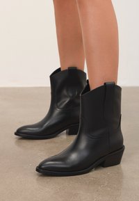 Botas occidentales de cuero negro hasta la mitad de la pantorrilla con puntas afiladas y tacones bajos, usadas sobre piernas despejadas en un suelo liso.