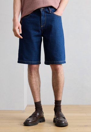 Homme portant un short en jean bleu foncé, des chaussettes marron et des mocassins marron foncé, debout sur un sol en bois contre un mur gris.