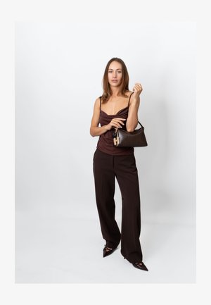 Bolso marrón con superficie texturizada, herrajes dorados y llavero con estampado. La modelo lleva un top y pantalones marrones en un entorno minimalista.