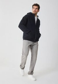 Marineblauwe hoodie met rits gemaakt van zacht materiaal, voorzien van een ritssluiting aan de voorkant, twee zijzakken en een capuchon met trekkoord, gecombineerd met lichtgrijze jeans en witte sneakers.