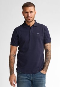 Námořnická polo košile vyrobená z bavlny, s klasikým límcem, krátkými rukávy a malým logem na hrudi. Nosená s modrými džíny.