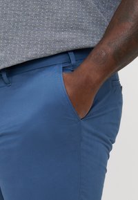 Siniset chinot, joissa on sileä pinta, sivutasku ja slim fit -design, yhdistettynä kuvioituun harmaaseen paitaaan.