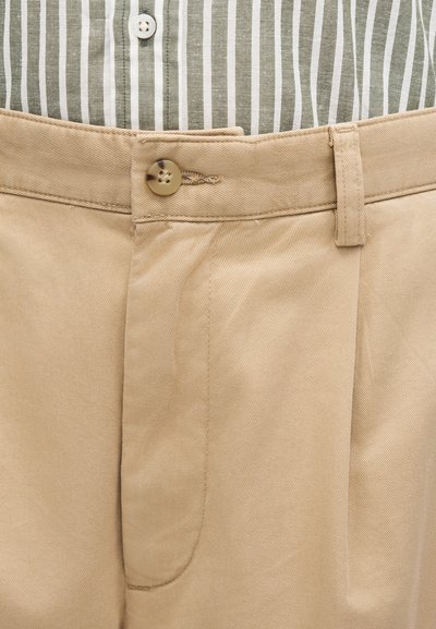 Pantaloni bej cu nasturi la talie, peste o cămașă în dungi verticale verde și alb, cu nasture alb vizibil.