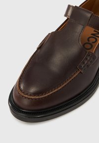 Κοντινό πλάνο σκούρου καφέ δερμάτινου μοκασίνι με λεπτομερή ραφές και μαύρη ελαστική σόλα σε λευκό φόντο.