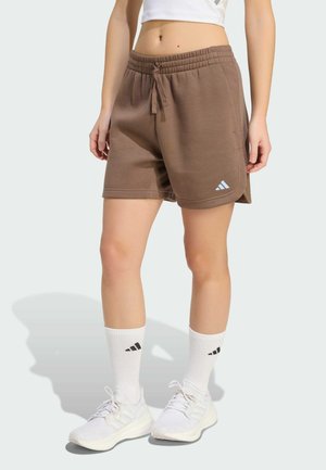 Person iført brune shorts med snøre, hvide crew-sokker med sort logo, hvide sneakers og en kort hvid top mod en lys baggrund.