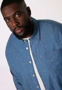 Blå chambrayskjorta med vita knappar och en ficka, med ett subtilt upprepat mönster, bärs över en vit t-shirt.