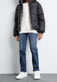 Chaqueta de plumas negra con detalles de logo, camiseta blanca, jeans de mezclilla azules y zapatillas blancas. Atuendo casual con diversas texturas y colores.