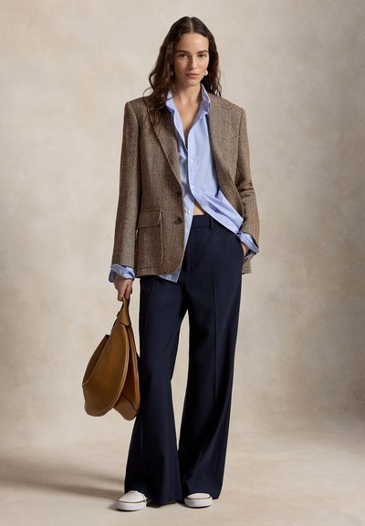 Woman wearing brown herringbone blazer over light blue shirt and wide-leg navy pants, holding large brown handbag, standing against neutral background.