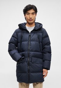 Tengerészkék pufikabát kapucnival, elülső cipzárral, két cipzáras mellzsebbel és egy nagy folt zsebbel az alsó elülső részen. Tűzött textúra.