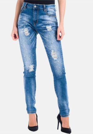 Vaqueros ajustados azul con parches desgastados y perlas decorativas dispersas, combinados con tacones altos negros.