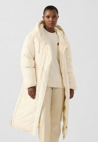 Lange crème pufferjas met een grote capuchon, voorzien van een ritssluiting aan de voorkant, zijzakken en een zachte, gewatteerde textuur. Draagt over een bijpassende outfit.