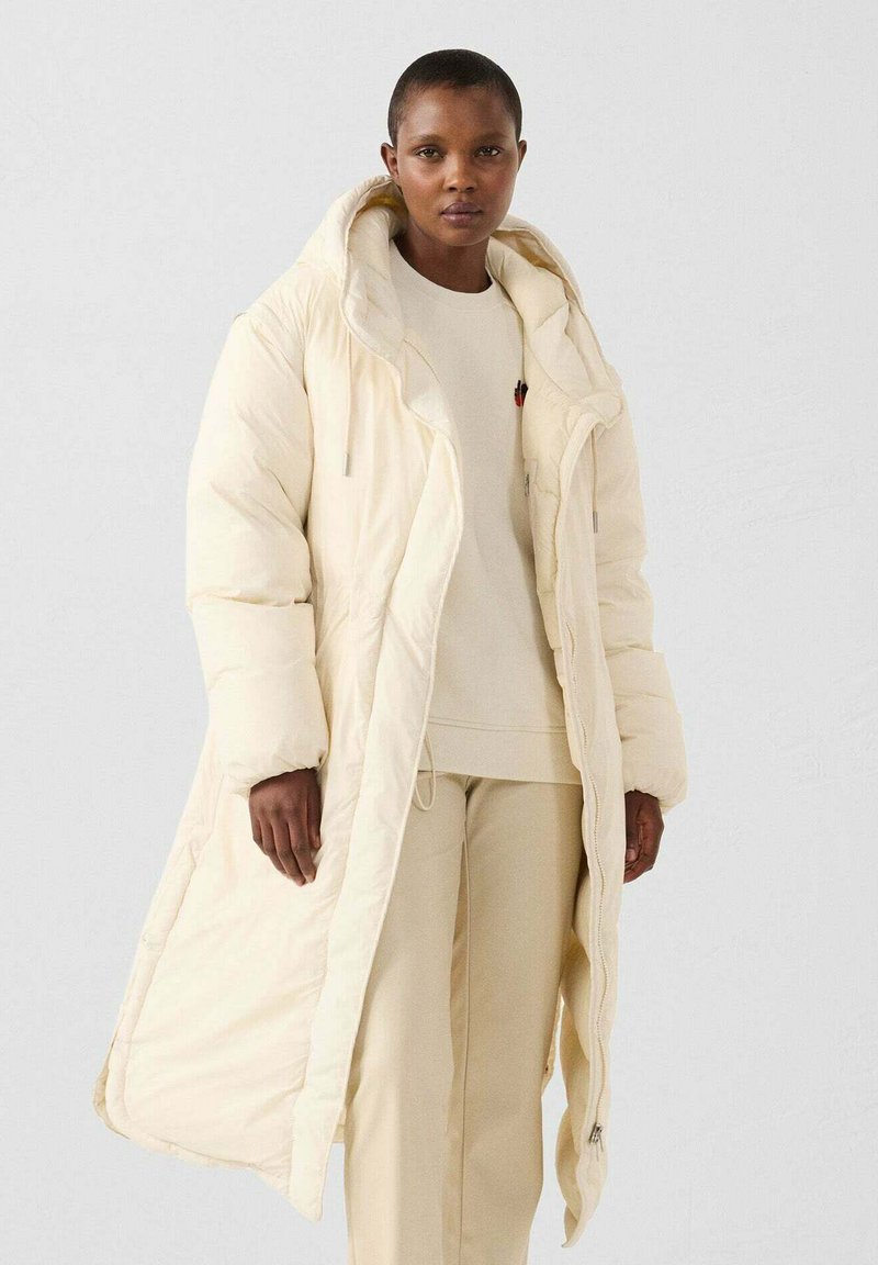 Lange crème pufferjas met een grote capuchon, voorzien van een ritssluiting aan de voorkant, zijzakken en een zachte, gewatteerde textuur. Draagt over een bijpassende outfit.