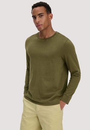 Mann trägt ein olivgrünes Langarmshirt mit Rundhalsausschnitt und beige Hose, steht mit den Händen in den Taschen vor hellem Hintergrund.