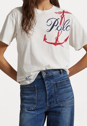 Frau trägt ein weißes T-Shirt mit einem großen roten Anker und dem Schriftzug „Polo“, kombiniert mit hoch taillierten blauen Jeans.