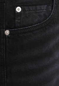 Černá džínová látka s hladkou strukturou, vybavená předním kapsou a stříbrnými kovovými detaily. Švy jsou viditelné podél okrajů.