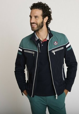 Homme aux cheveux bouclés foncés et à la barbe, portant une veste zippée bleu sarcelle et bleu marine par-dessus un polo bleu marine, souriant, mains dans les poches du pantalon bleu sarcelle, regardant vers la gauche.