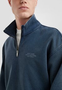 Dunkelblauer, zip-in Sweatshirt mit hohem Kragen. Auf der Brust befindet sich weißer Text: "EGALE WAS, NUR ENSPANEN." Weicher Stoff.