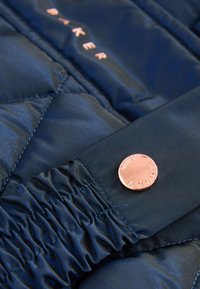 Marineblauwe doorgestikte jas van glanzend materiaal, met gerimpelde manchetten en een ronde metalen knop met de gegraveerde merknaam.