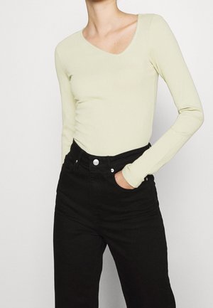 Persona que lleva puesto un top de canalé amarillo pálido de manga larga metido en pantalones negros de cintura alta con una mano en el bolsillo.