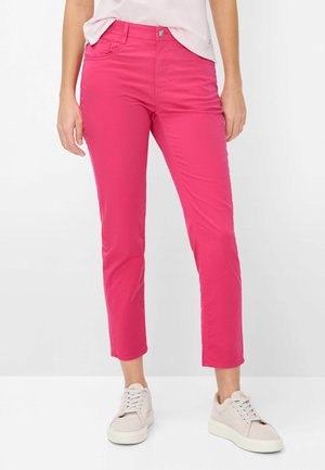 Femme portant un pantalon slim rose vif jusqu'à la cheville et des baskets à lacets rose clair, debout devant un fond blanc uni.