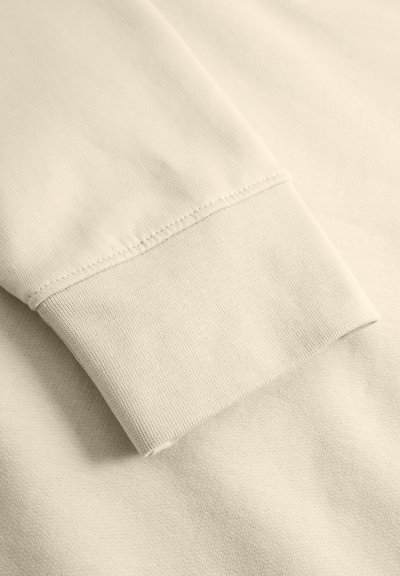 Le poignet du sweatshirt en coton beige clair présente une texture côtelée avec des coutures soignées. Le tissu doux a une finition lisse, replié sur le bord.