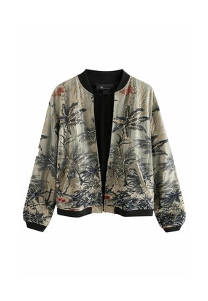 Femme portant une veste bomber à motif floral et une longue jupe sombre, debout pieds nus sur une rive rocheuse avec des vagues de l'océan au coucher du soleil.