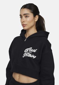 Felpa corta nera in tessuto morbido, con la scritta grafica bianca "Good For Nothing" sul davanti. Zip frontale e dettagli con cappuccio.