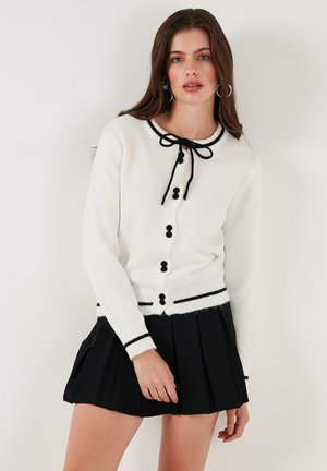 White knit cardigan with black accents, round neckline, black buttons, and a bow tie at the neck. Worn with a pleated black skirt.