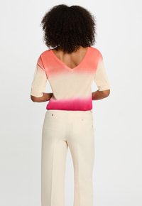 V-hals, kortärmige trui in verloopkleuren van koraal naar roze, gecombineerd met lichtbeige broek met twee achterzakken en een gladde structuur.