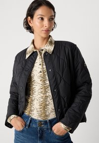 Donna che indossa un giubbotto nera trapuntato sopra una camicia beige a righe di tigre con colletto e jeans blu, con le mani in tasca, che guarda di lato.