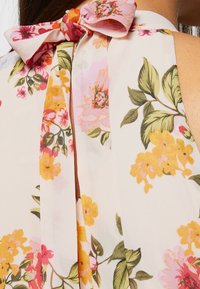 Gros plan sur un vêtement à col licou fleuri présentant des fleurs roses, jaunes et oranges avec des feuilles vertes et un nœud noué à l'arrière.