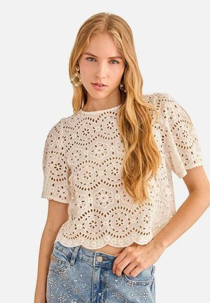 Joven mujer con pelo largo y rubio lleva grandes pendientes, un top blanco de encaje calado y jeans azules con adornos de perlas, posando contra fondo blanco.