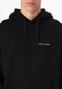 Homme portant un sweat à capuche noir avec une poche avant et un petit texte blanc "STYLE & SCOTT" sur la poitrine, sans autres détails visibles.