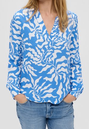 Frau trägt eine blau-weiß gemusterte Bluse mit langen Ärmeln und V-Ausschnitt, Hände in den Taschen und hellblaue Jeans.