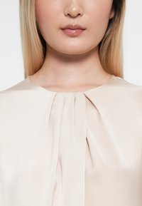 Béžová saténová blůza s kulatým výstřihem a jemnými sklady na přední straně, která ukazuje hladkou strukturu a decentní design.
