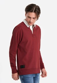 Bordeauxfarbenes Sweatshirt mit weißem Kragen, langen Ärmeln, strukturiertem Stoff und einem markanten schwarzen Etikett auf der unteren linken Seite.