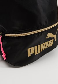Sac Puma noir avec fermeture éclair dorée et logo, avec un cordon de tirette rose visible sur un fond clair.