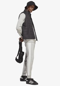 Črn podložen brezrokavnik preko sivega hoodija, sive trenirke z belimi črtami, črn klobuk s širokim robom ter črne športne copate z belimi detajli.