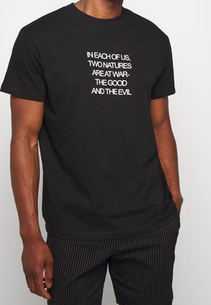 Maglietta nera in cotone con testo bianco: "DENTRO OGNUNO DI NOI, DUE NATURE SONO IN GUERRA: IL BENE E IL MALE." Maniche corte, scollatura rotonda.
