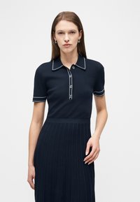 Γυναίκα που φοράει φόρεμα πόλο κοντομάνικο σε ναυτικό μπλε με λευκές ραφές στο γιακά και τα μανίκια, στέκεται μπροστά σε απλό λευκό φόντο.